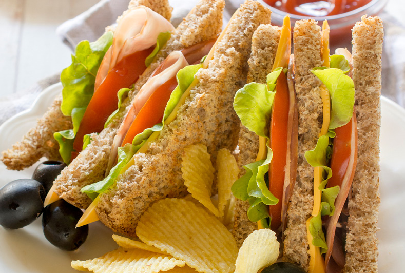 close up photo of turkey sandwhich with lettuce tomato cheese on toasted wheat bread with chips and black olives on the side