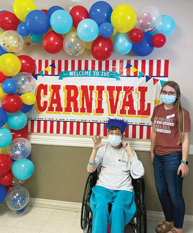 Irvine staff member standing next to resident Janet R. in front of CARNIVAL banner and balloons